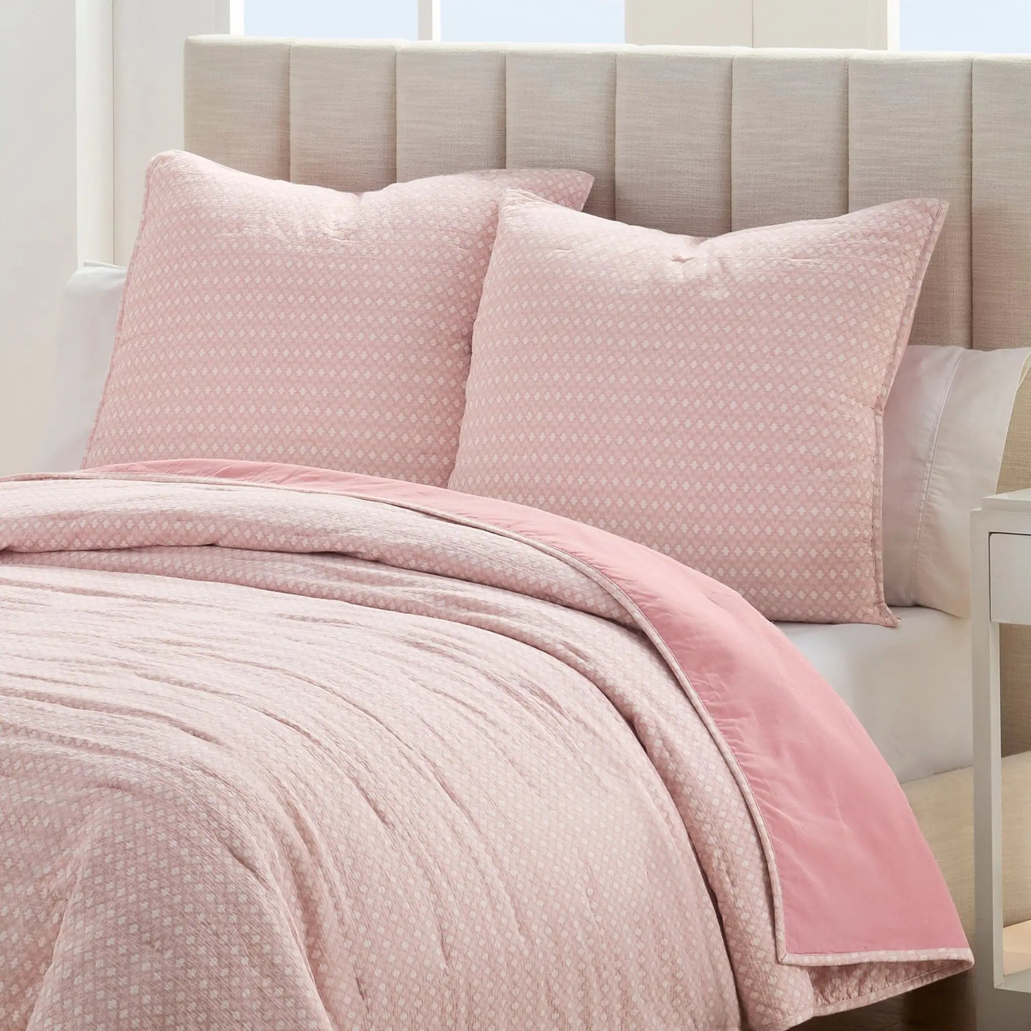 A pink quilt with a subtle white geometric pattern, featuring two matching euros styled on a bed.