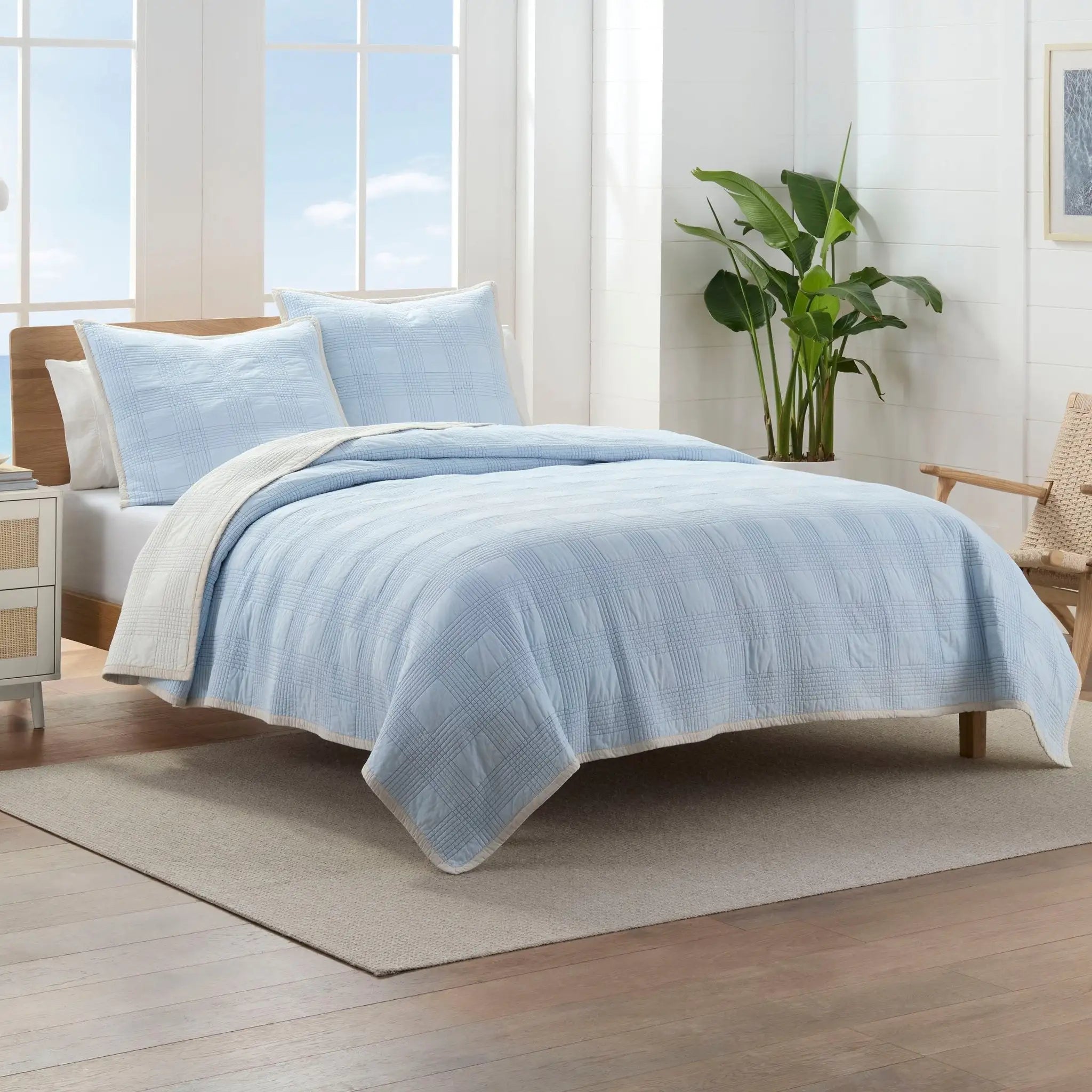 Bed with light blue quilt and euros in a bright bedroom with large windows.