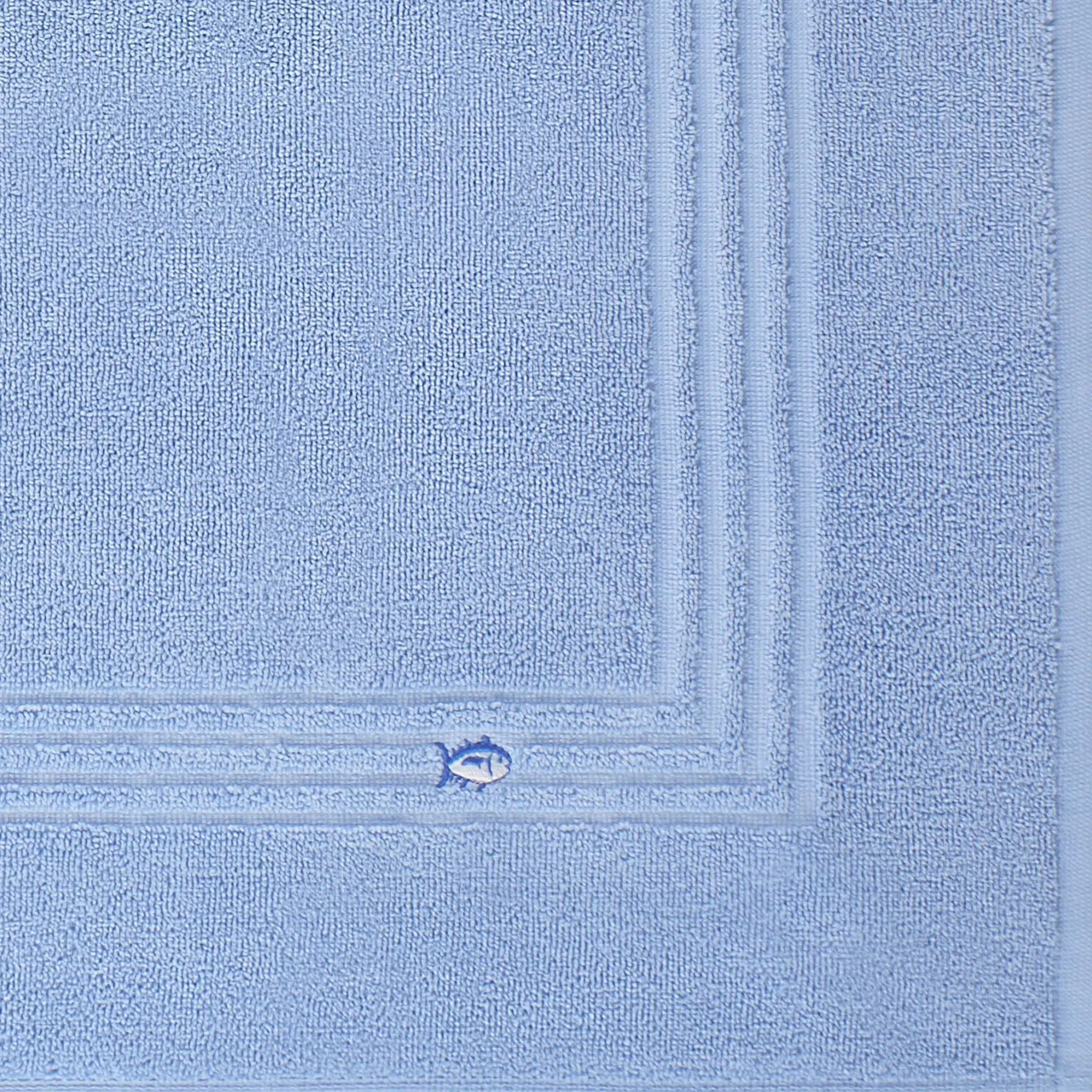 A close-up of a blue textured bath mat fabric with a stitched border and a small embroidered fish motif near the corner.