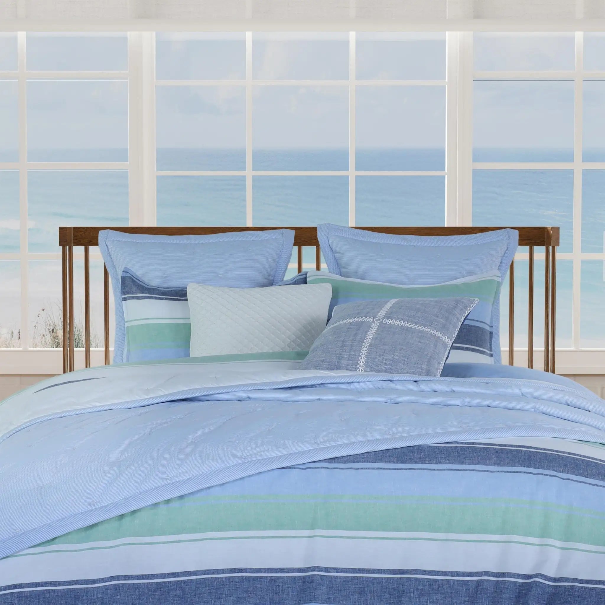 A bed dressed in a striped bedding set with light blue, navy, mint green, and white stripes, styled with matching pillows in front of a large window showing a beach view.