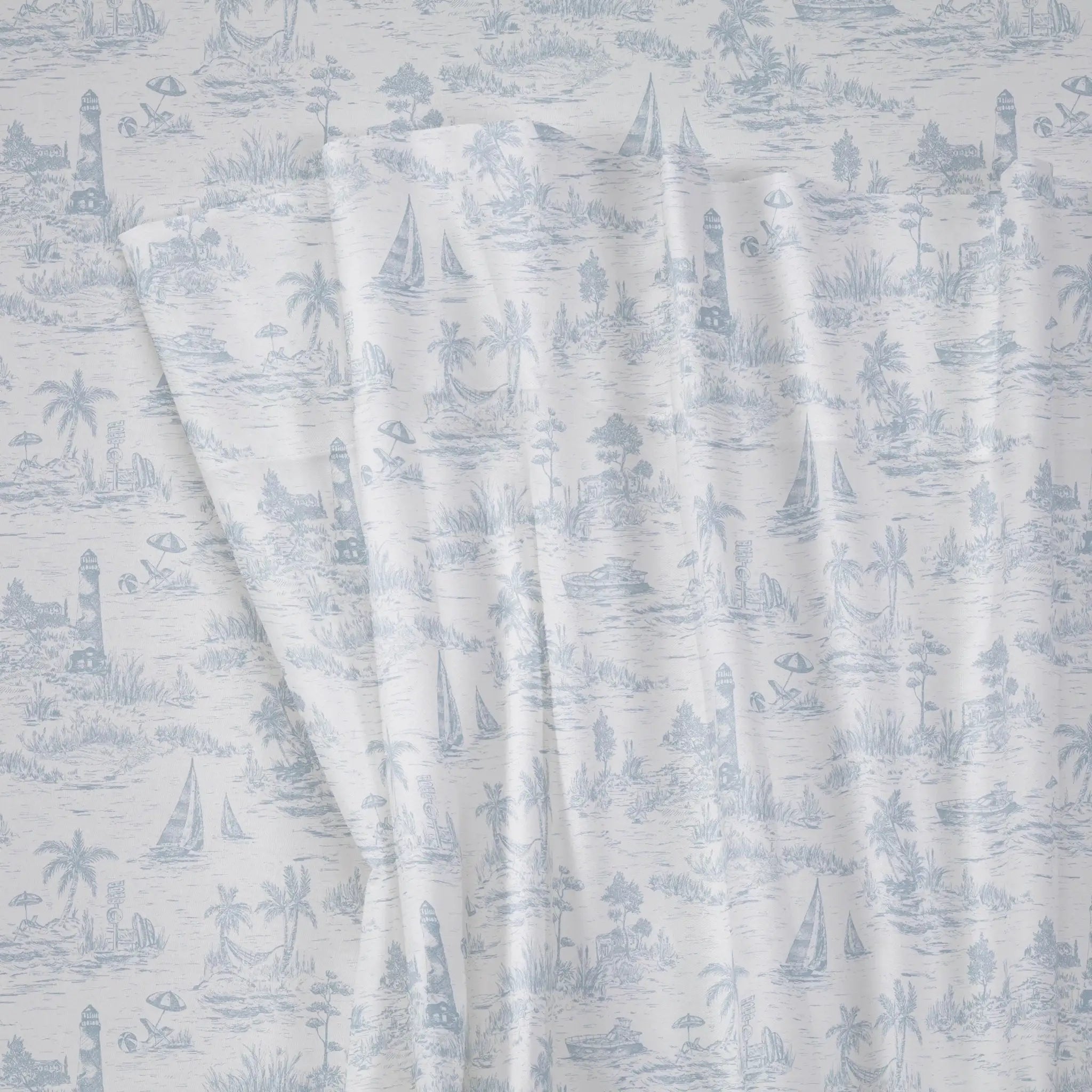 A close-up of a fabric with a light blue toile de Jouy pattern on a white background, showing draped folds and a nautical scene with sailboats, palm trees, and lighthouses.