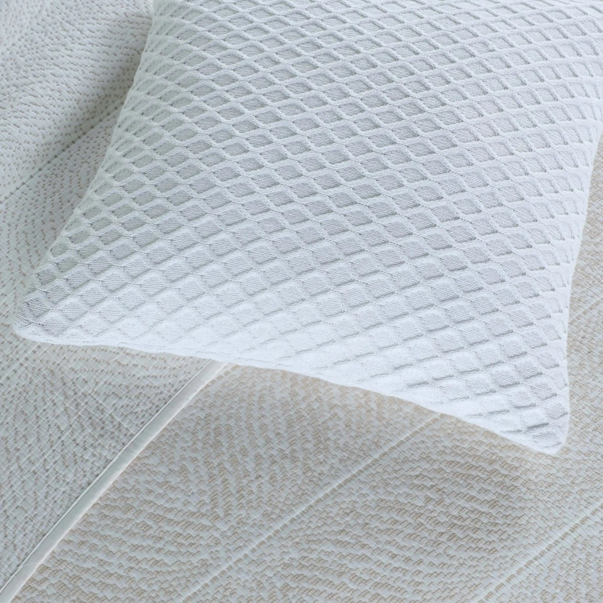 A close-up of a white textured pillow with a diamond-patterned cover resting on a matching white quilted bedsheet.