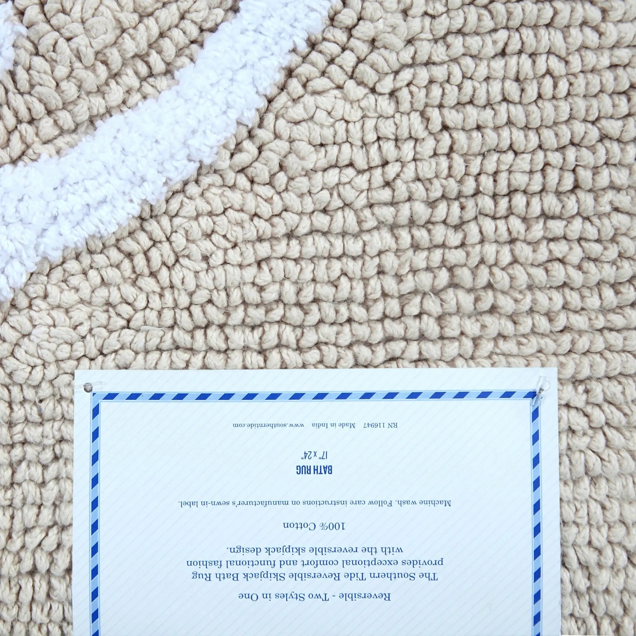 A close-up of a sand textured rug with a white stripe, featuring a white paper label pinned near the bottom with blue border lines and small product details.
