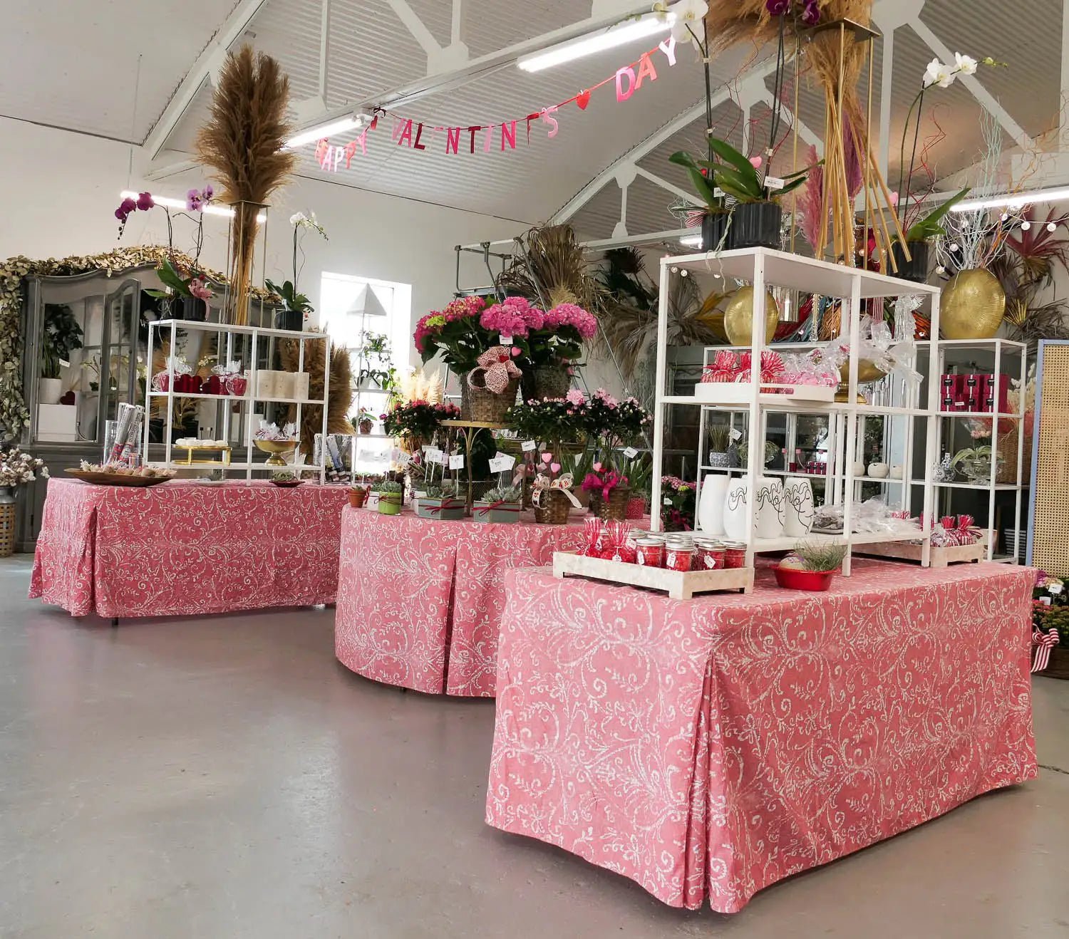 A Valentine's Day themed event setup in a warehouse or studio space, featuring three tables draped in washed red patterned tablecloths.