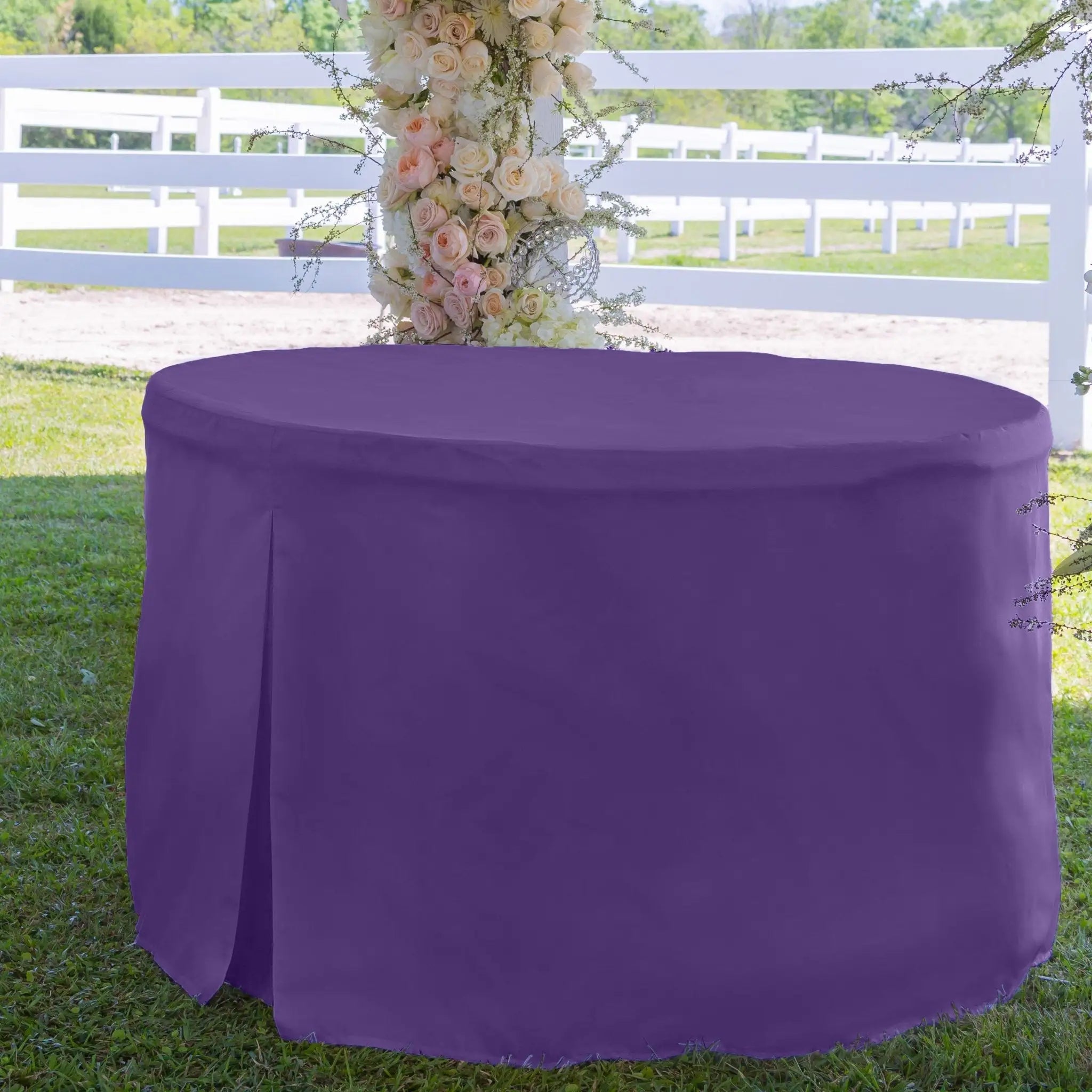 Violet color tablecloth on a round table with floral decorations against a white fence and greenery background