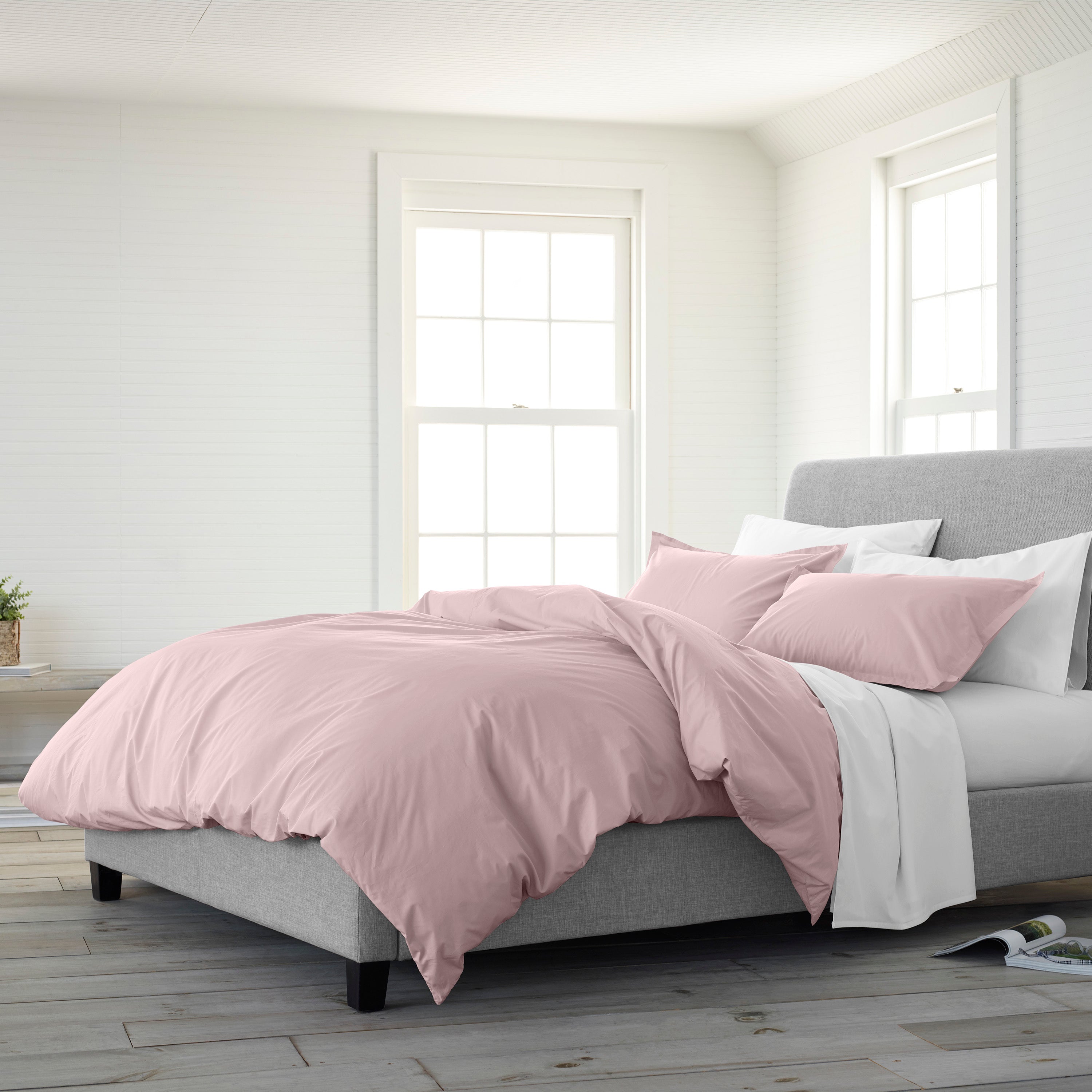 Light pink duvet set on a gray bed with white and pink pillows, in a bright, airy room with large windows.