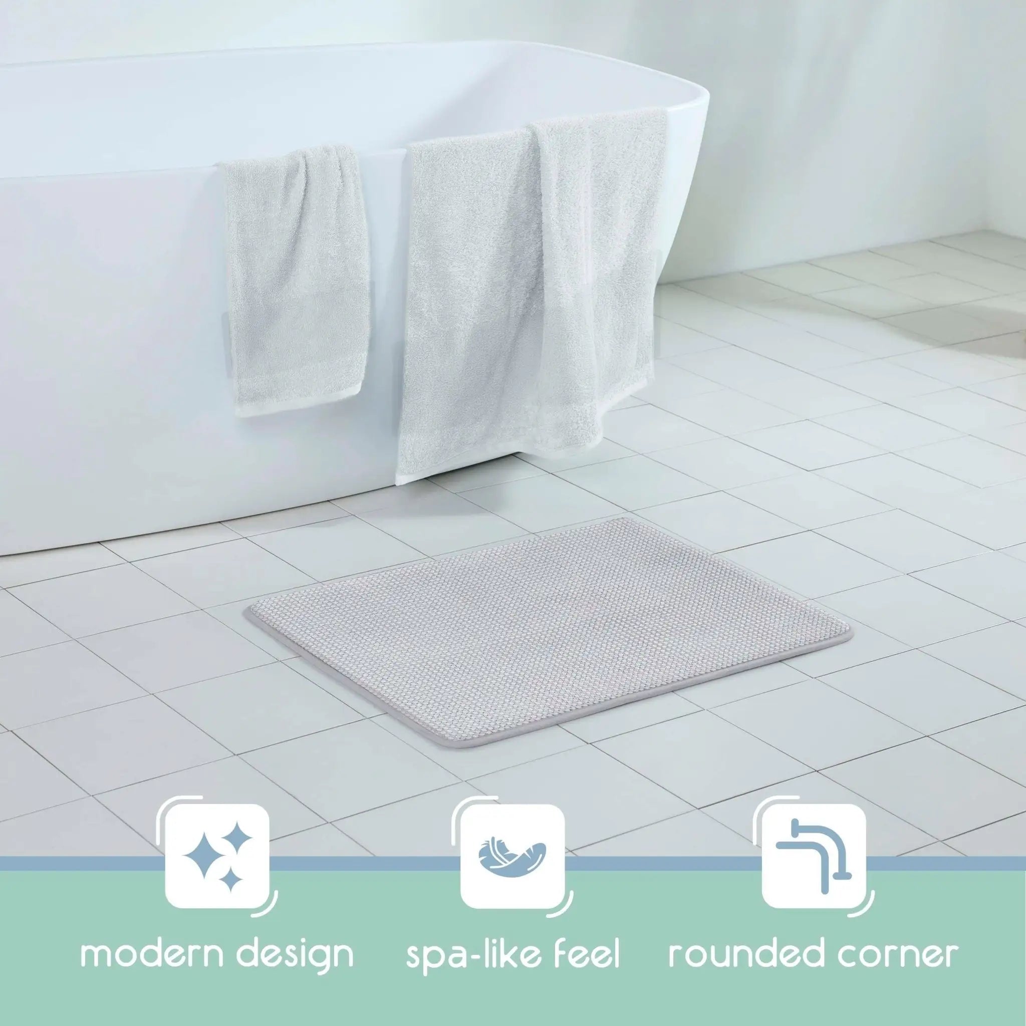 A light gray textured bath rug with rounded corners placed on a white tiled bathroom floor next to a white freestanding bathtub with white towels draped over the edge. Three feature icons and text at the bottom read: modern design, spa-like feel, and rounded corner.