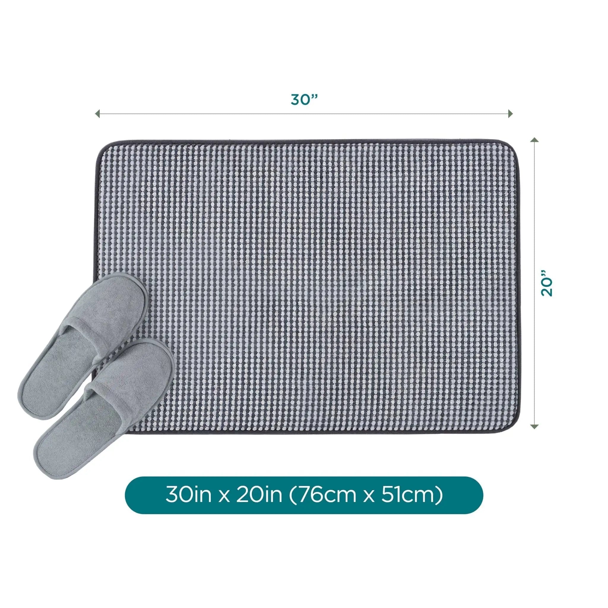 A rectangular black bath rug shown with a pair of gray slippers on a white background, with dimension arrows labeling it 30inches x 20 inches (76 x 51 cm) and a teal text bar at the bottom.