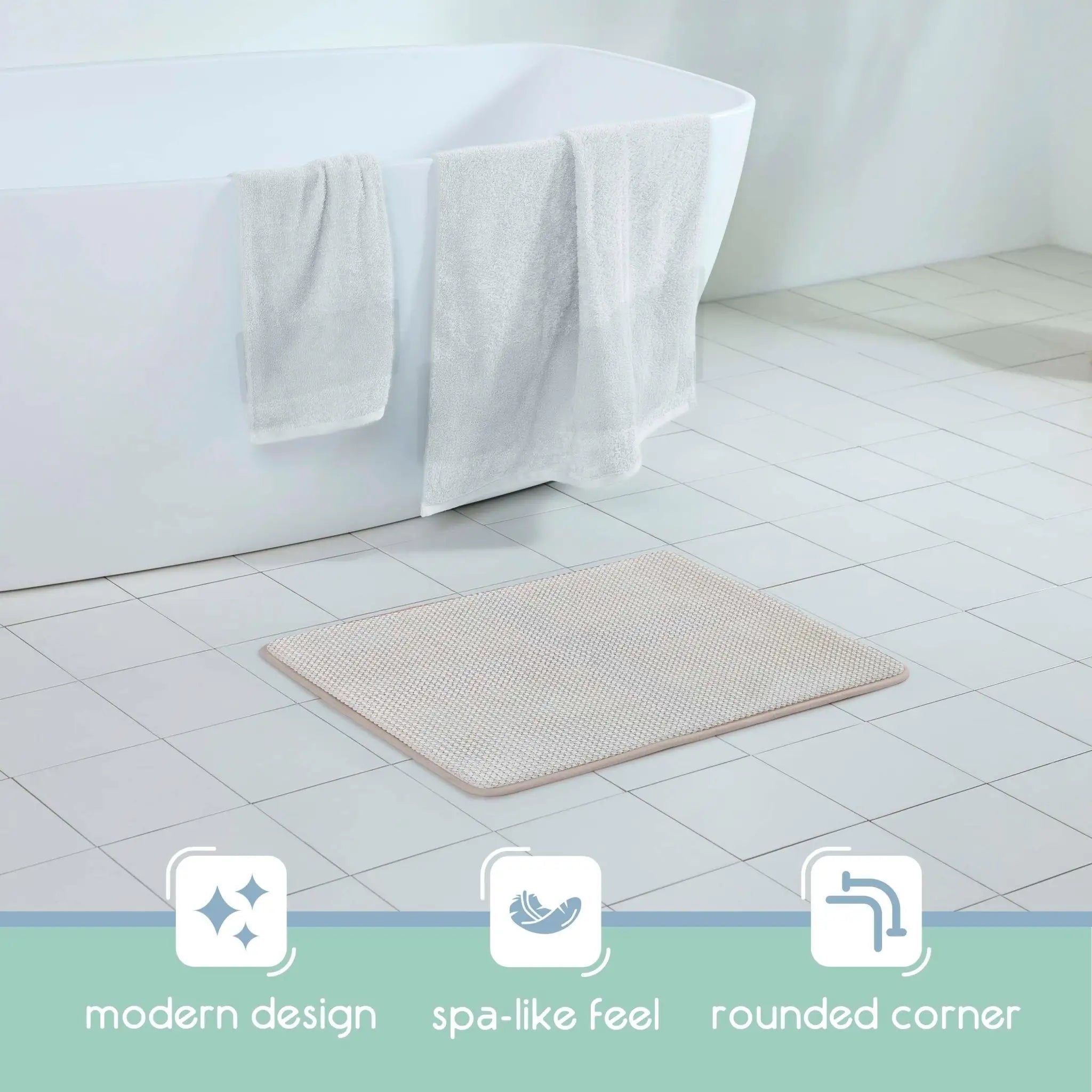 A rectangular sand bath rug with rounded corners, placed on a white tiled bathroom floor next to a white bathtub with white towels draped over the edge. Three feature icons and labels are shown along the bottom: 'modern design', 'spa-like feel', and 'rounded corner'.