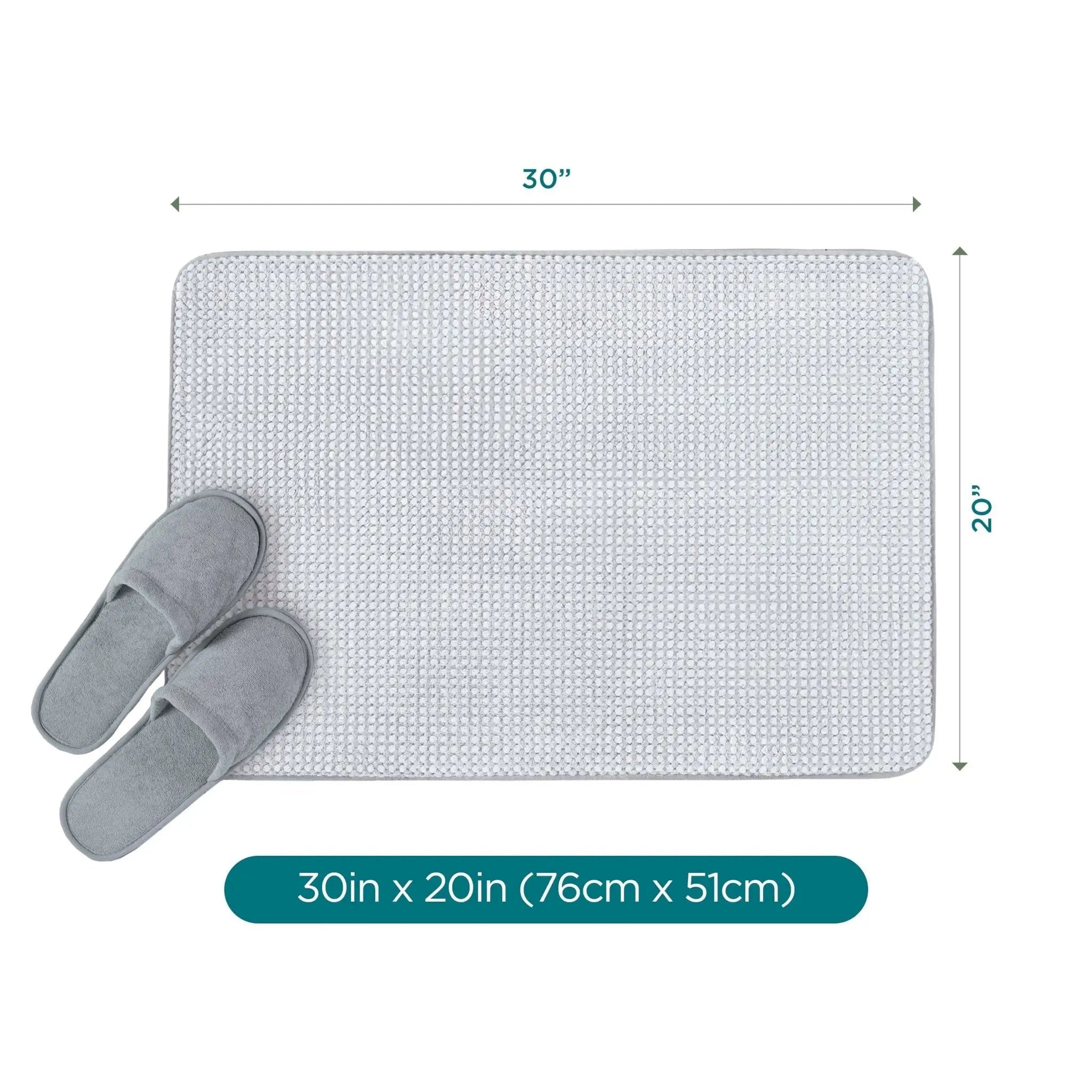 A rectangular light gray bath rug with rounded corners shown on a white background, with dimension arrows indicating 30 inches by 20 inches. A pair of gray slippers rests on the lower left, and a teal label at the bottom reads 30in x 20in (76cm x 51cm).