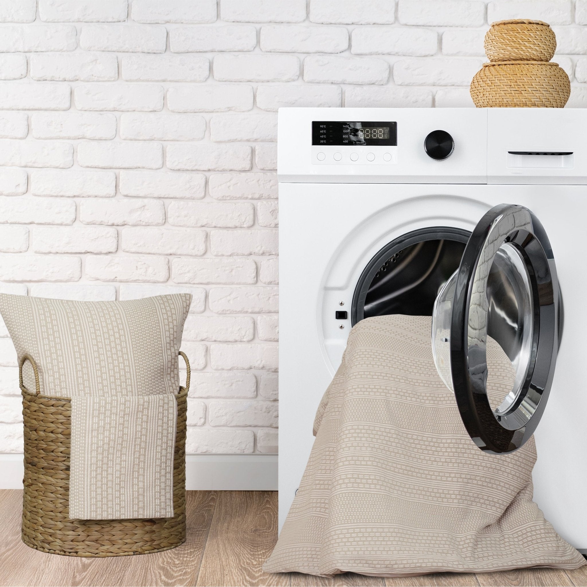 Washing machine with open door, beige comforter, and laundry basket against a white brick wall.
