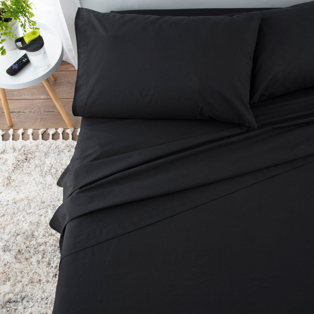 Black sheet set with two pillows on a bed, accompanied by a small table with a plant and remote control.