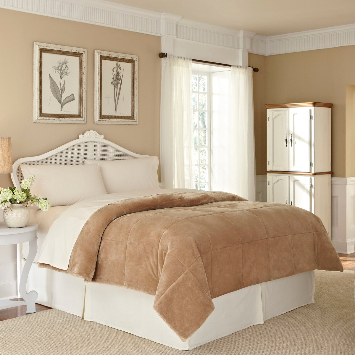 Tan plush blanket draped over a bed with white linens in a softly lit room featuring floral artwork and a cabinet.