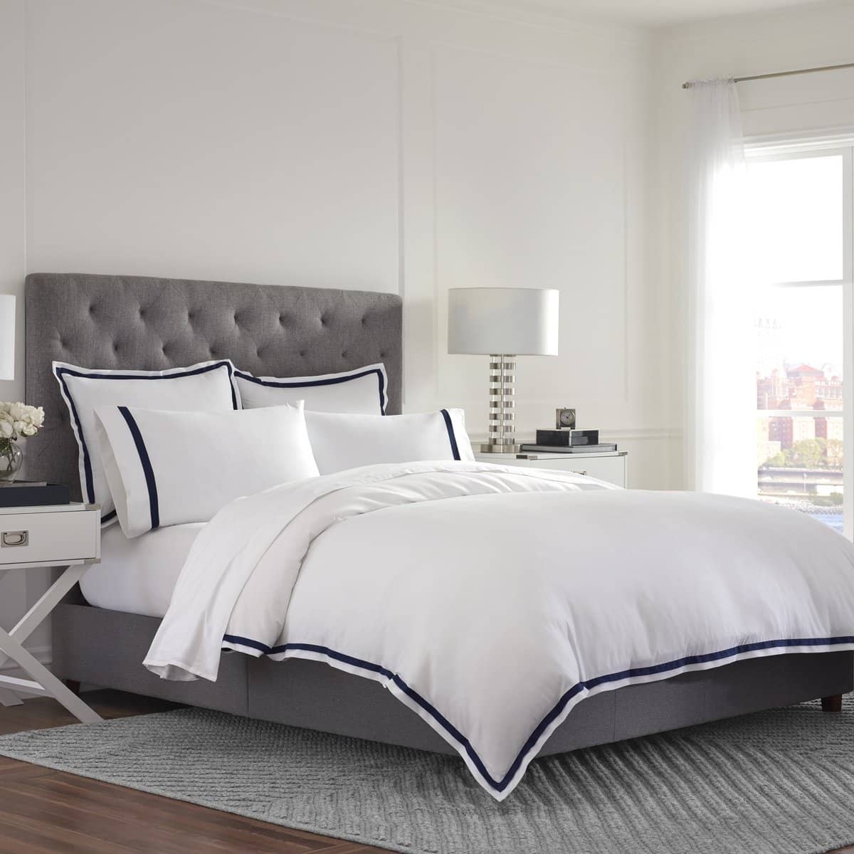 White duvet cover set with navy trim, paired with matching pillow shams, on a gray upholstered bed.