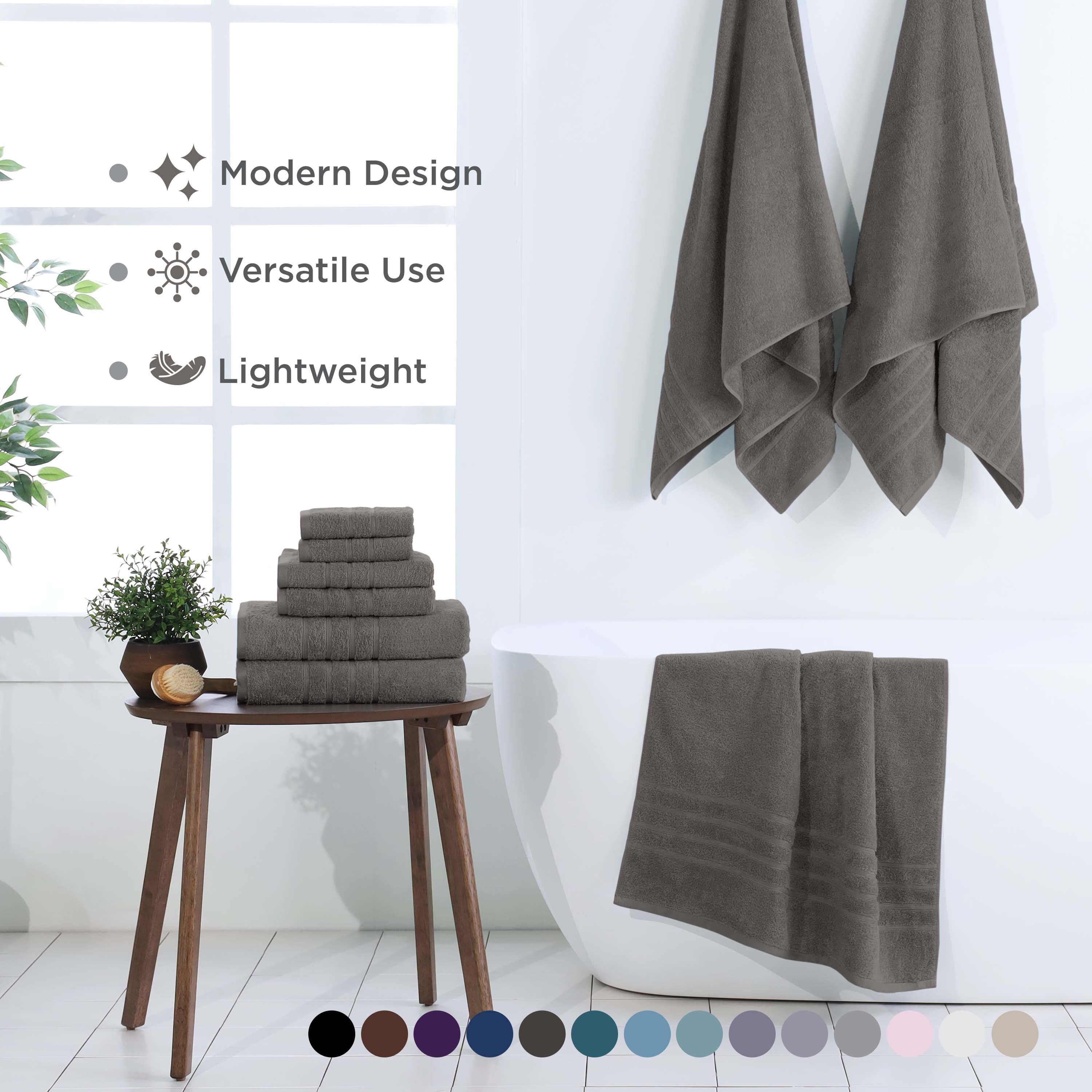 Six gray towels stacked on a wooden table with two hanging on a wall near a bathtub and a plant in the background.