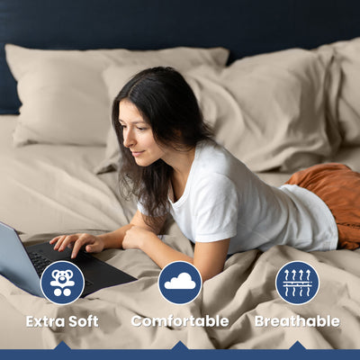 A woman is lying on a bed with beige sheets, using a laptop and looking focused.