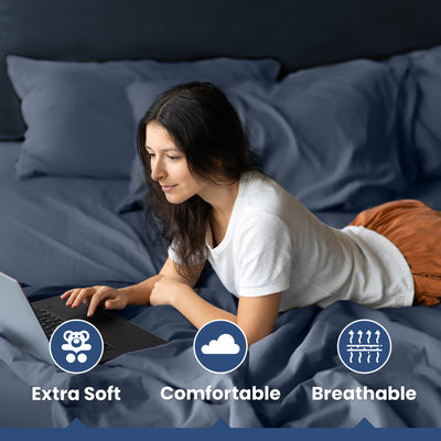 A woman is using a laptop while lying on a bed with dark blue cotton sheets and pillows.