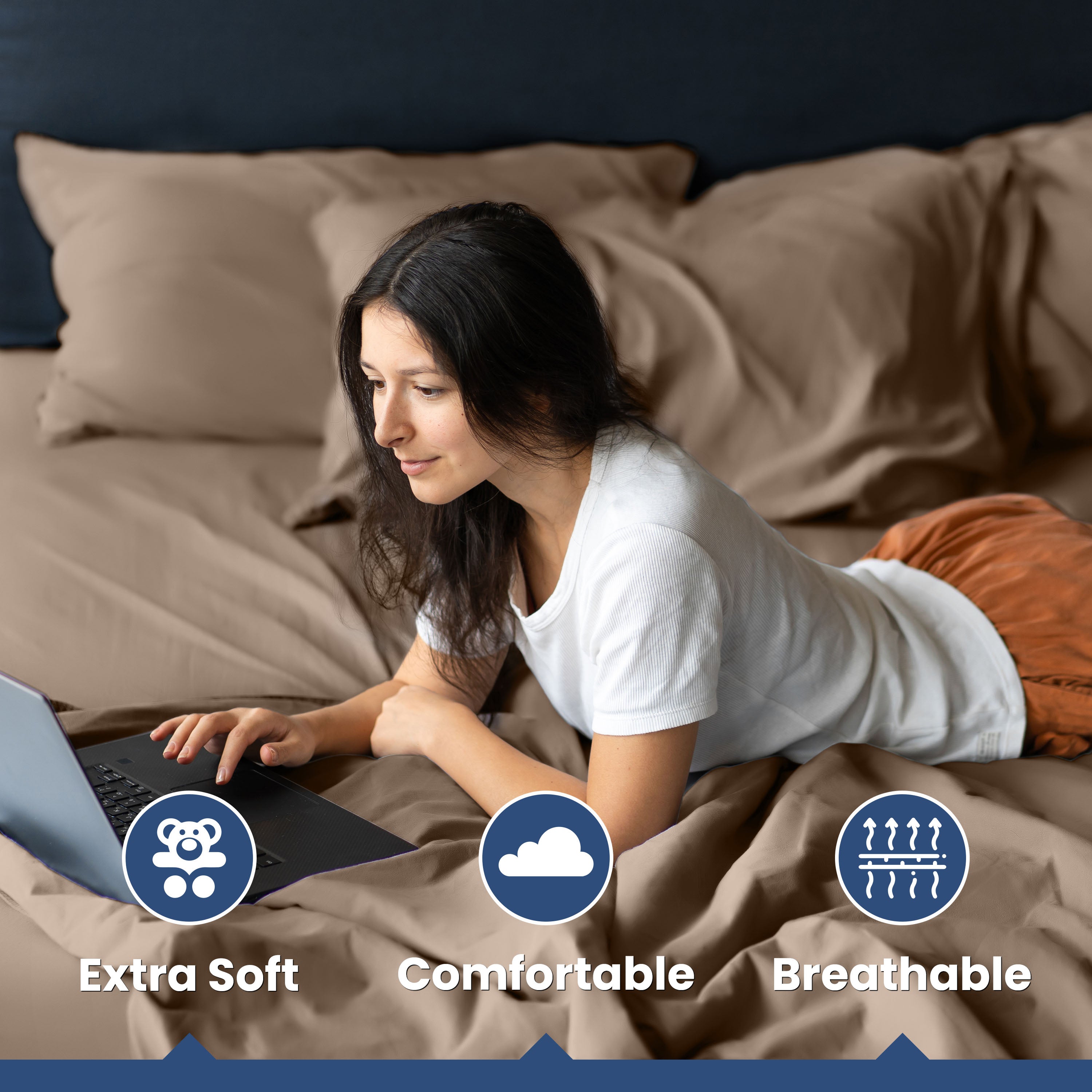 A woman in a white shirt uses a laptop while lying on a bed with brown sheets and pillows.