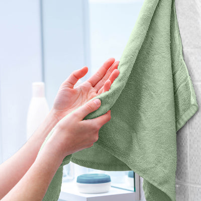 Hands holding a soft green towel hanging on a wall, with bathroom products visible in the background.