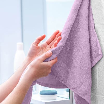 Hands holding a soft, lavender towel hanging on a bathroom wall with blurred toiletries in the background.