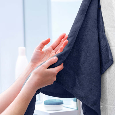 Hands holding a dark navy towel hanging on a bathroom wall with toiletries in the background.