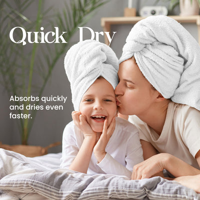 Two people, an adult and a child, wearing white towels on their heads, smiling and sitting on a bed with bedding.