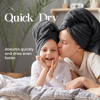 A woman kisses a smiling child, both wearing dark towels on their heads, sitting on a bed with light bedding.