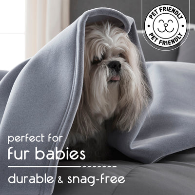 A small dog with long fur is partially covered by a gray blanket on a couch.