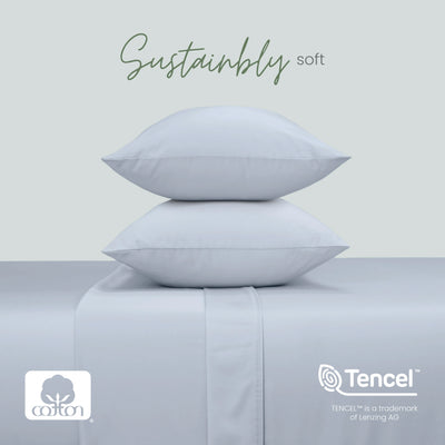 Two blue pillows stacked on a matching upholstered surface, with the words 'Sustainably soft' above. Cotton and Tencel logos appear at the bottom, with a pale gray background.