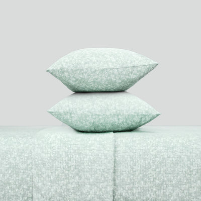 Two decorative pillows in a soft green pattern stacked on a matching sheet set.