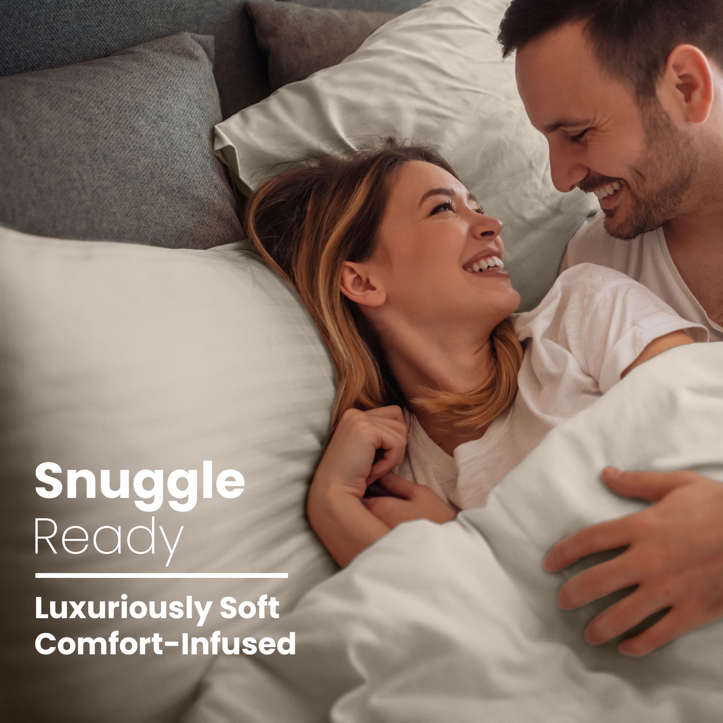 Couple smiling and cuddling in bed with light-colored pillows and sheets.