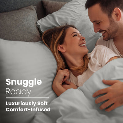 Couple smiling and cuddling in bed with light blue cotton sheets and pillows.