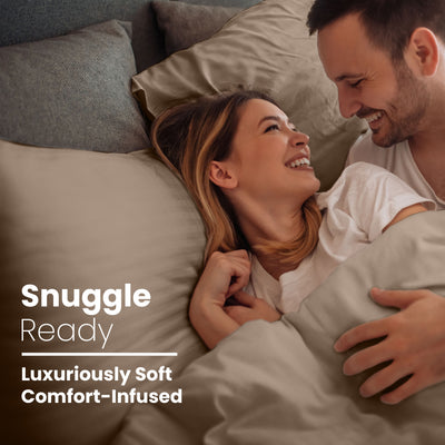 Couple smiling and cuddling in bed with soft, neutral-colored sheets and pillows.
