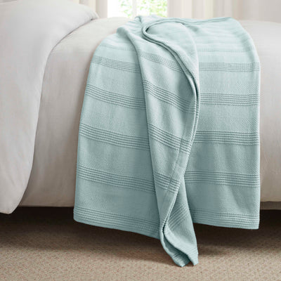 Light blue cotton blanket with textured stripes draped over the edge of a bed.