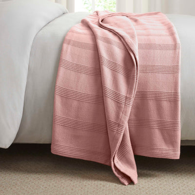Cotton blanket in a soft pink color with textured stripes, draped over the edge of a bed.