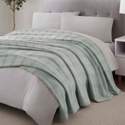 Light green cotton blanket draped over a bed with gray bedding and pillows, next to a nightstand.