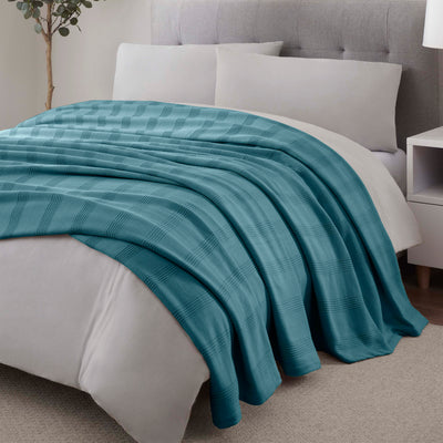 Teal cotton blanket with a textured plaid pattern draped over a bed with white pillows and a light gray headboard.