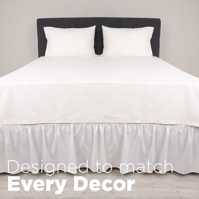 White ruffle bed skirt drapes over the edge of a bed with two white pillows on a gray headboard.