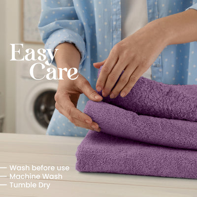 Hands folding purple towels on a table, with a washing machine visible in the background.