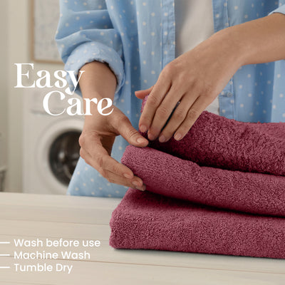 Hands folding stacked burgundy towels on a light surface, with a washing machine visible in the background.