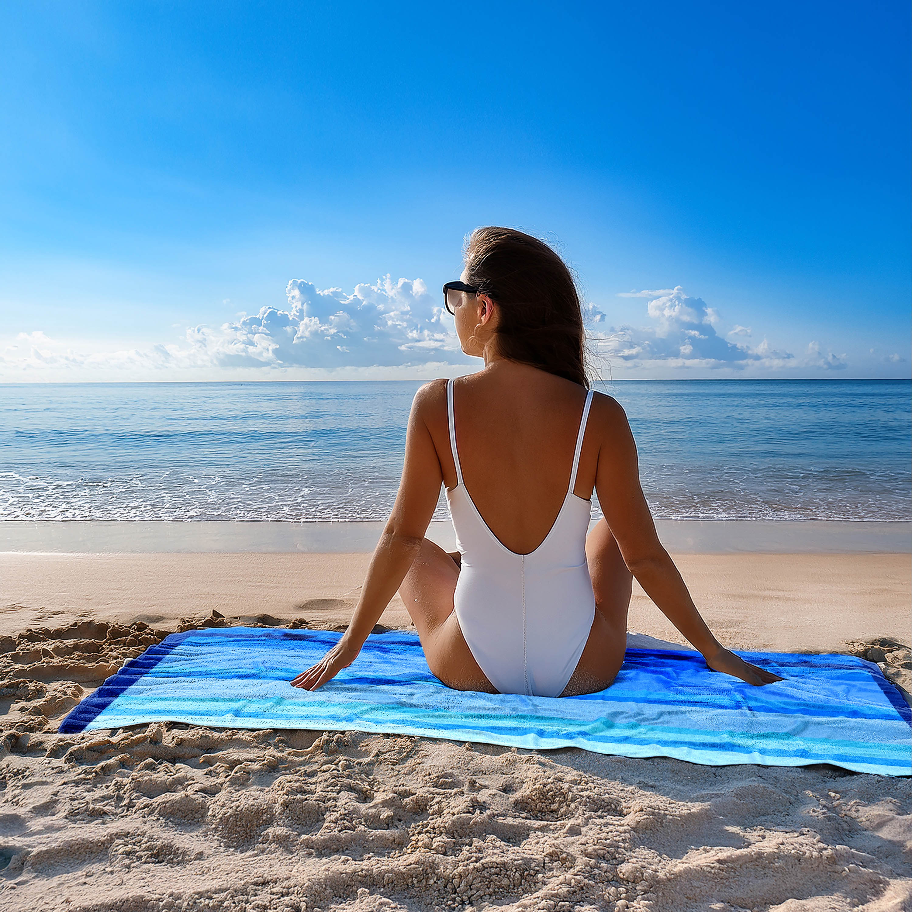 Woman in a white swimsuit sitting on a blue towel at the beach with a clear sky.