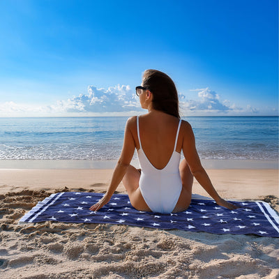 Woman in a white swimsuit sitting on a blue towel at the beach with a clear sky.