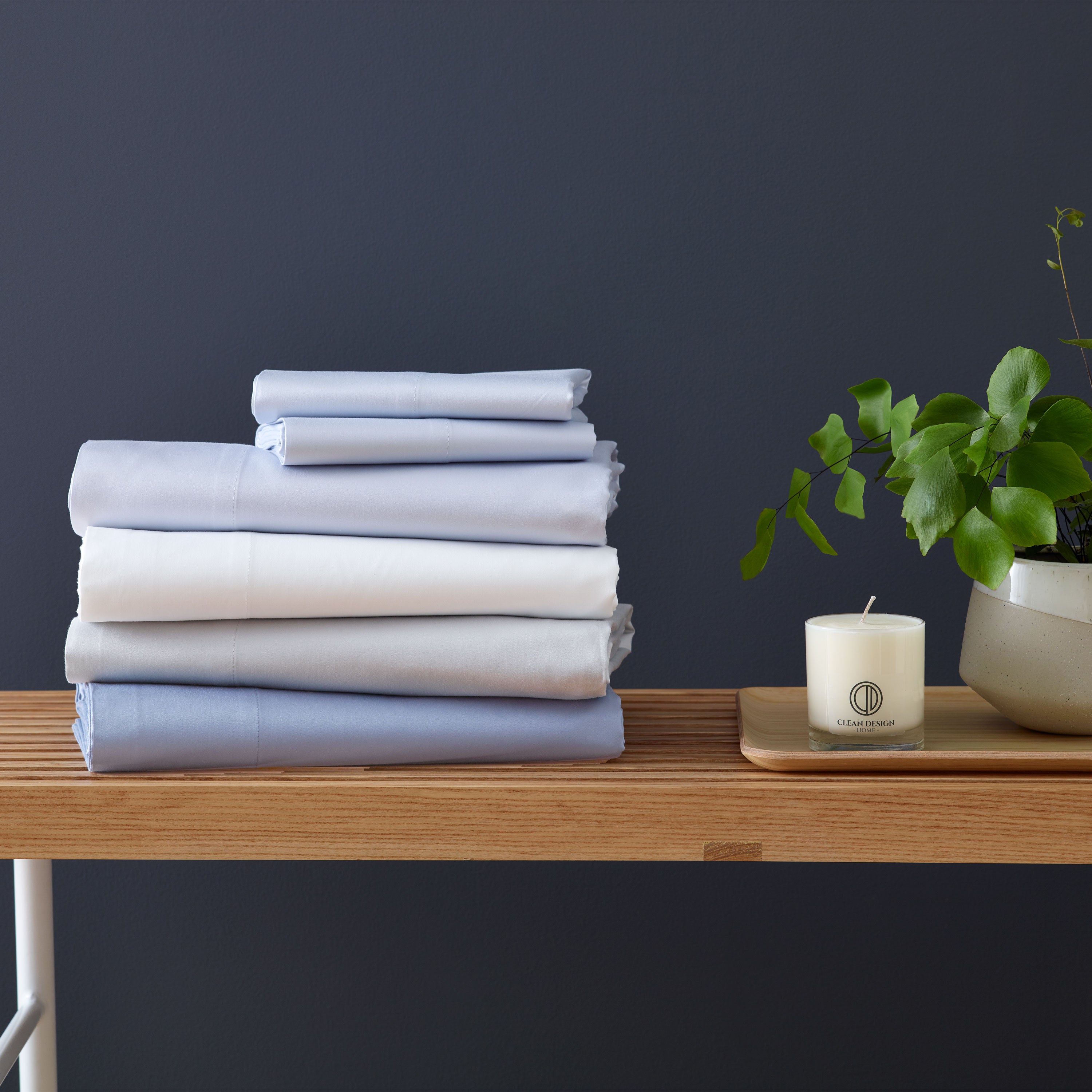 Stack of light blue and white sheets on a wooden shelf next to a candle and a small potted plant.