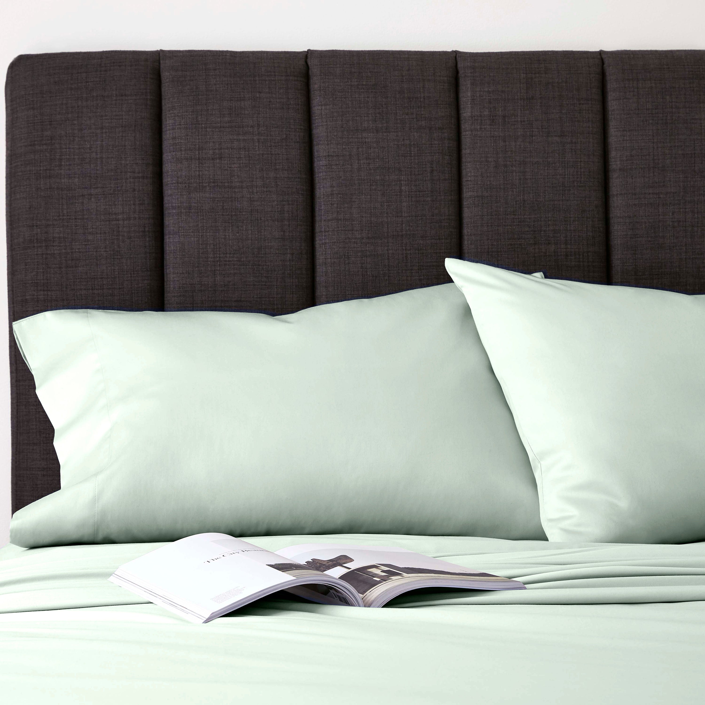 Light green pillowcases on a bed with a dark gray headboard and an open magazine on the sheets.