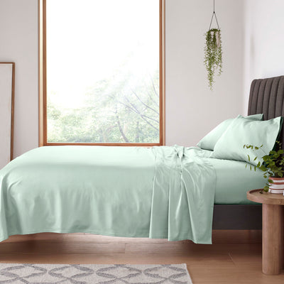 Light green sheet set laid on a bed with two matching pillows, beside a wooden side table and a window.