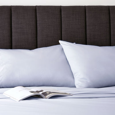 Light blue sheet set with two pillows on a bed, next to an open magazine and a dark upholstered headboard.