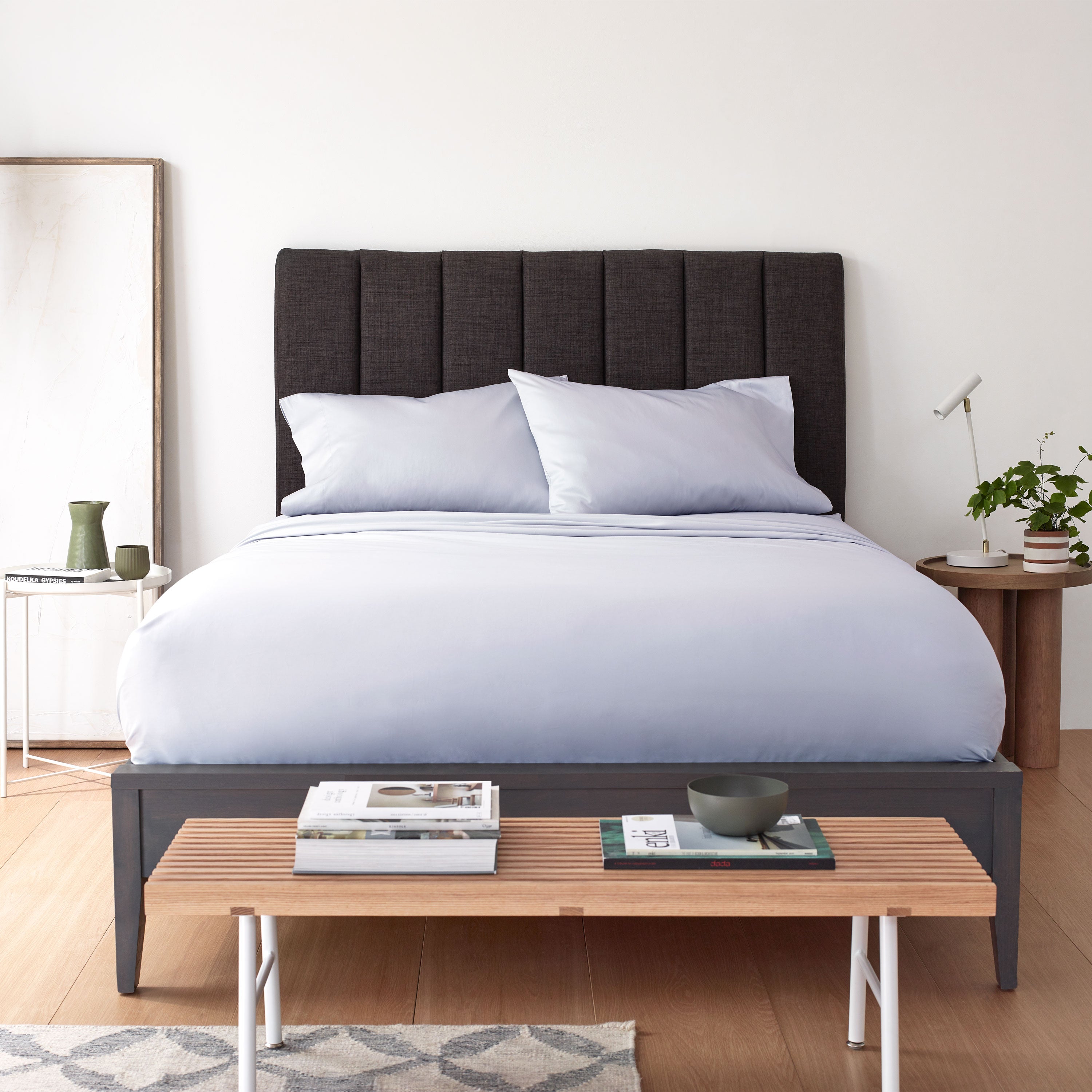 Light blue sheet set on a bed with a dark headboard, accompanied by a side table and a wooden bench with books.