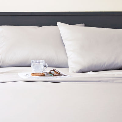 Light gray sheet set with two pillows, a glass of water, a coaster, and sunglasses on a magazine.