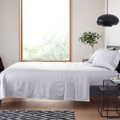 Light gray sheet set on a bed with two pillows, a black pendant light, and a plant near a large window.
