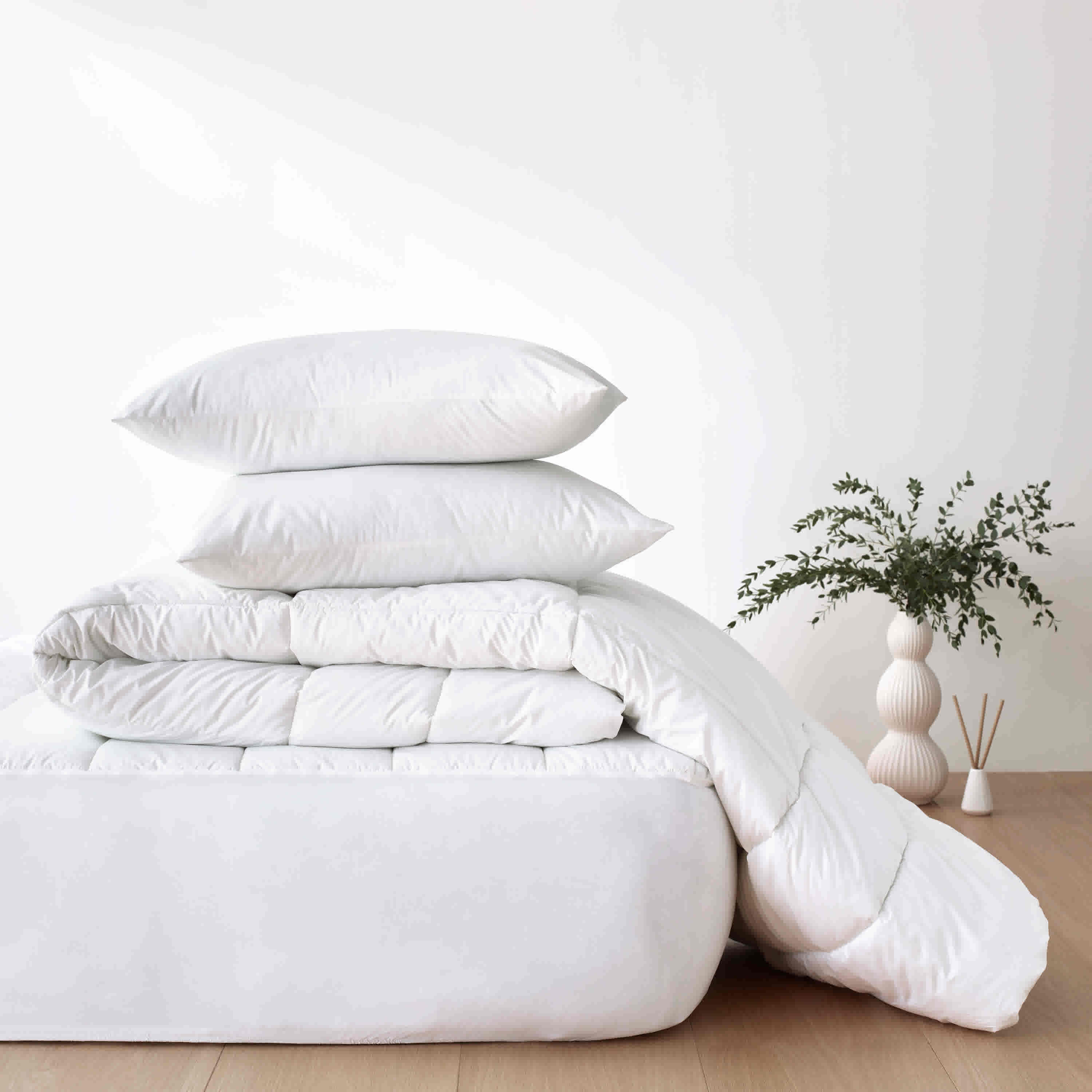 Stack of white pillows and a comforter on a mattress pad, with a decorative vase and plant in the background.